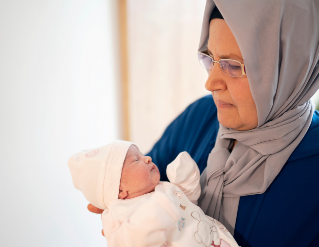 an older woman cradling a newborn baby. The woman, wearing a light gray hijab and glasses, gazes lovingly at the infant, highlighting a profound sense of connection and care. The baby, swaddled in a soft, cream-colored outfit adorned with subtle patterns, wears a matching hat that emphasizes its delicate features. 